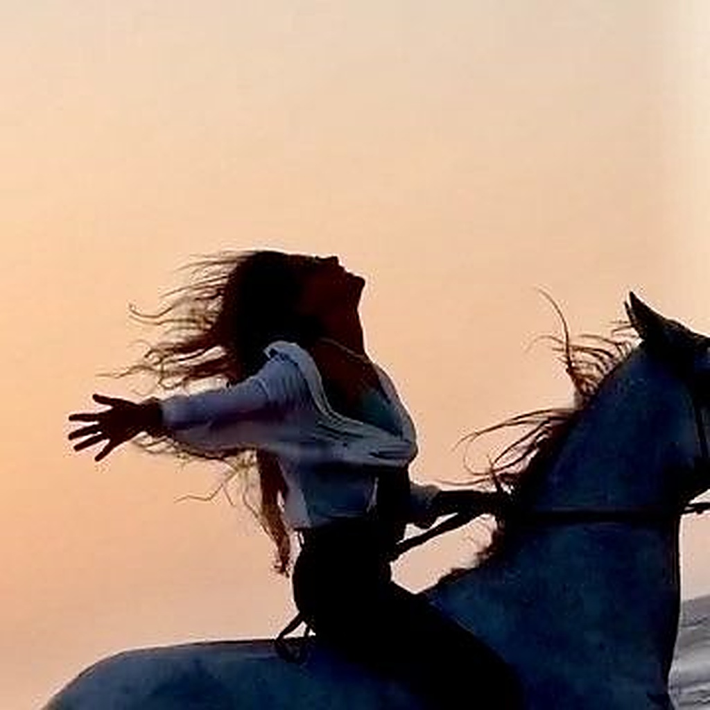 A woman riding a horse at dusk, head thrown back and arms open